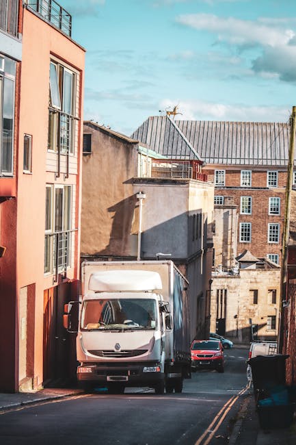 A white moving van parked on a narrow street in an urban residential area with multi-storey buildings on either side. The van is positioned close to the left sidewalk, partially blocking the entrance to a building with a red brick facade and large glass windows. The street slopes downward and is lined with parked cars, including a small red car behind the van and a dark vehicle further down. In the background, there are older brick buildings with flat roofs, some featuring rooftop antennas. The scene is illuminated by natural daylight, with shadows cast by the buildings. This setting illustrates the logistical challenges of home relocation and furniture transport in tight access urban environments, typical of logistics handled by Removal Van Catford during people’s moves or packing and moving operations.