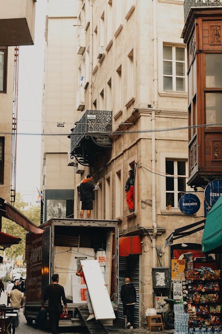A scene of a home relocation process on a narrow urban street in Catford, with a removals vehicle parked close to a historic stone building featuring multiple windows and a decorative balcony. A crew member wearing a black jacket and orange helmet is standing on an extended platform lifting or securing items inside the building. Nearby, another person in a red jacket assists with the process. The truck's side door is open, revealing packing materials such as cardboard boxes, plastic-wrapped furniture, and packing blankets, ready for loading. The street is lined with shops, including a newsstand with snacks and drinks, and the scene is lit by natural daylight, showing an active moving operation that likely involves careful packing and furniture transport as part of a residential moving service by Removal Van Catford. Items are being carefully carried through the building’s doorway onto the vehicle, emphasizing the logistics of tight access jobs during a house or apartment removal in the area.