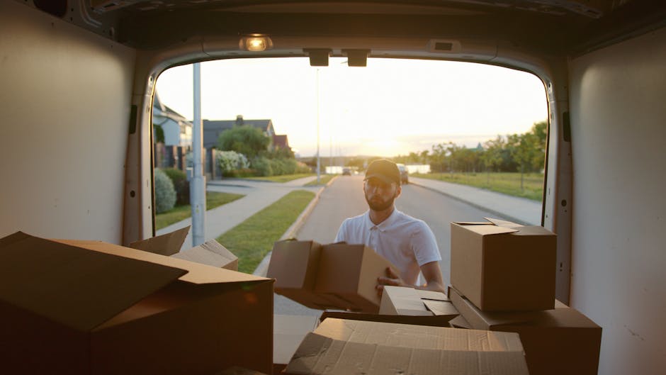 A man wearing a white polo shirt and sunglasses is loading medium-sized cardboard boxes into the back of a moving van parked on a residential street during sunset. The interior of the van shows severa