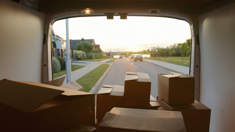 A man wearing a white polo shirt and sunglasses is loading medium-sized cardboard boxes into the back of a moving van parked on a residential street during sunset. The interior of the van shows severa