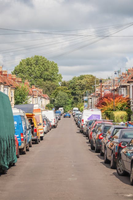 A residential street scene showing a wide, straight pavement lined with parked cars on both sides, including hatchbacks, sedans, and vans, with some covered by protective tarps and others with open doors; in the background, there are brick terraced houses with front gardens, trees, and shrubs, under an overcast sky with grey clouds. The image captures part of the loading area where Removal Van Catford might position their vehicle during a house move, with an emphasis on parking considerations and the environment for home relocation or furniture transport in Catford, as discussed in the Mountsfield Park small move removals insider tips page.