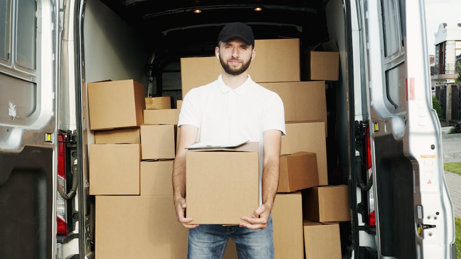 A man dressed in a blue jacket, dark trousers, and white sneakers is in the process of loading cardboard boxes into the open rear of a white removal van parked on a cobblestone street in an urban area